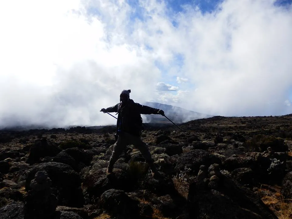 Silhouette of a Kilimanjaro guide with trekking poles walking across a volcanic rock field beneath dramatic clouds