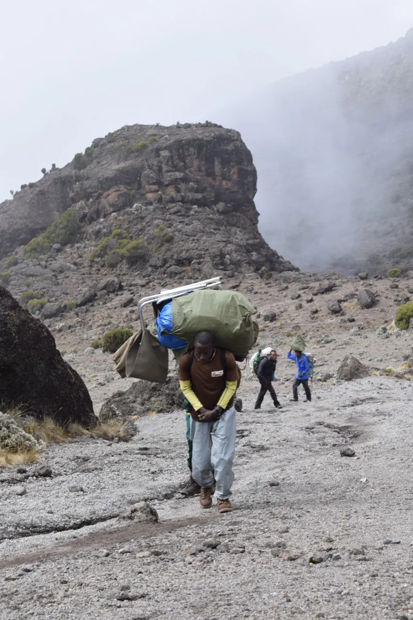 A porter carrying large duffel bags and baskets up a rocky trail on Mount Kilimanjaro, mist rising behind him